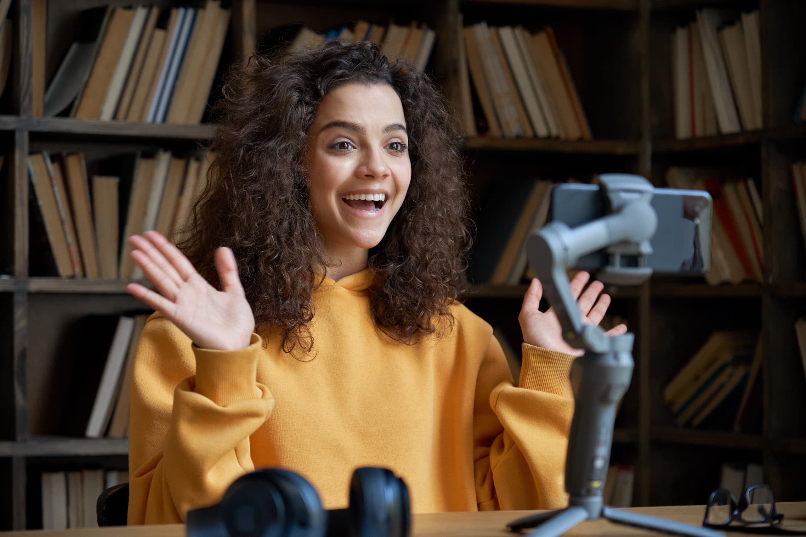 Excited hispanic teen girl social media channel blogger recording vlog on digital smartphone cam in library. School student vlogger talking looking at mobile phone on tripod shooting blog, streaming.